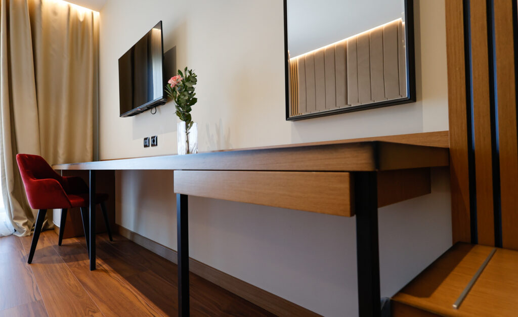 Long oak desk with red velvet chair, wall-mounted flat-screen TV and a mirror reflecting the headboard