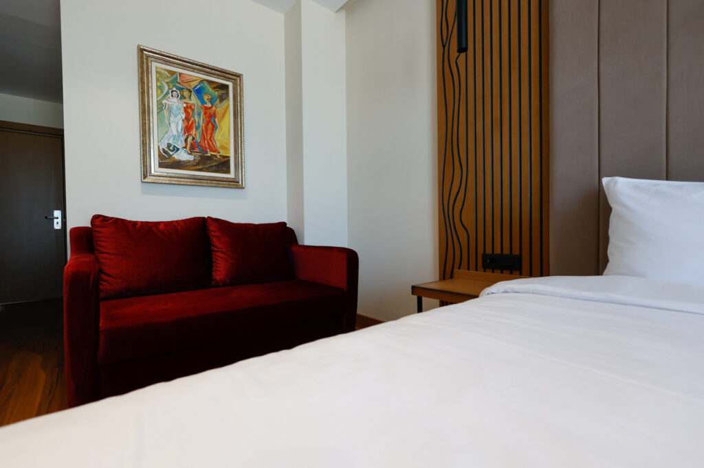 Red velvet sofa under a framed painting beside the bed and slatted oak panel, Valza Suite