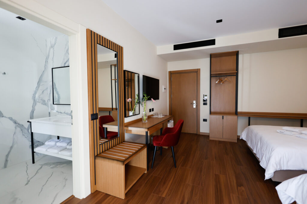 Double Twin Room with open marble-effect bathroom, desk, red chair, wardrobe and a bed with folded towels