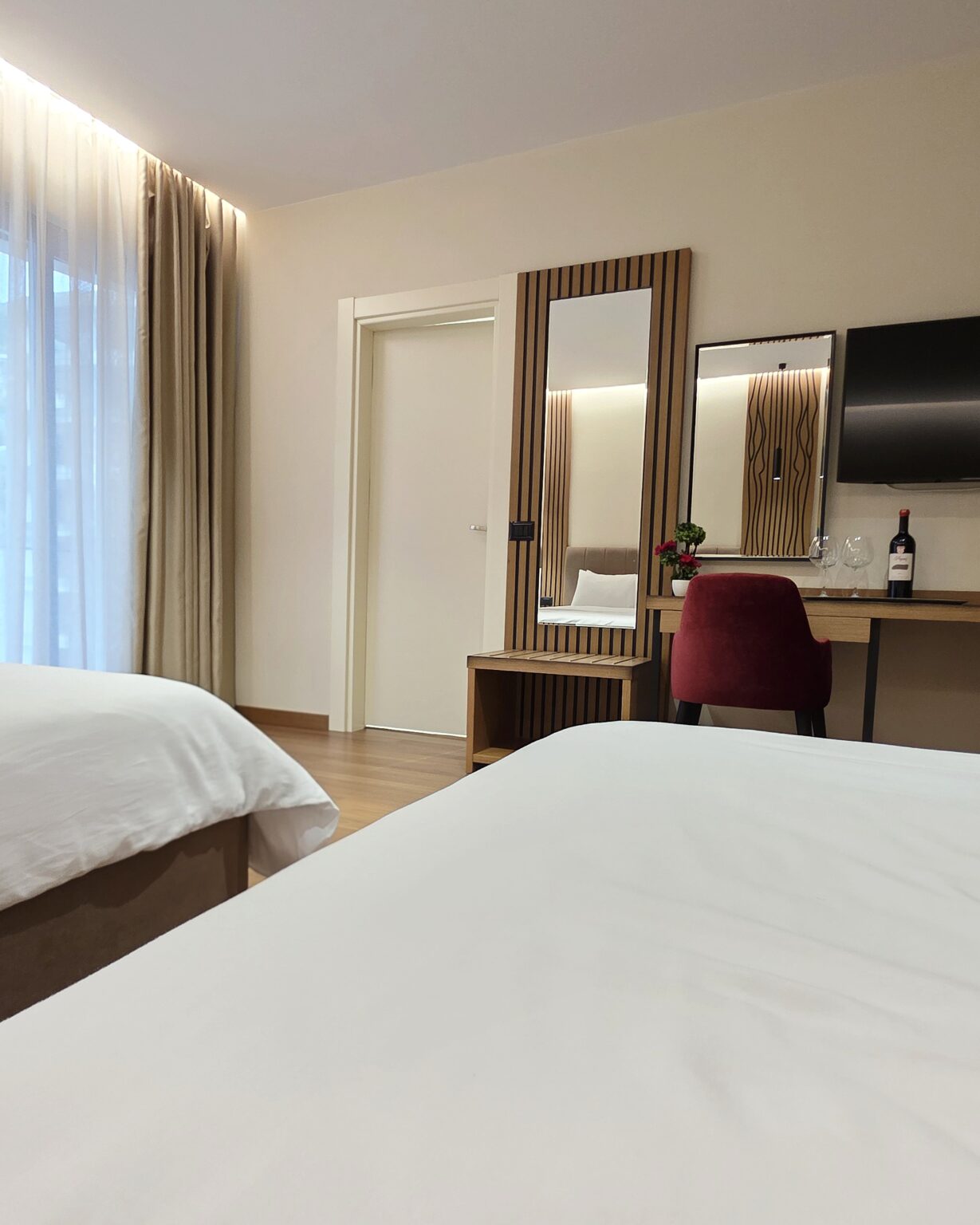 Double Twin Room seen from the beds, with desk, red chair, wall-mounted TV and wine bottle