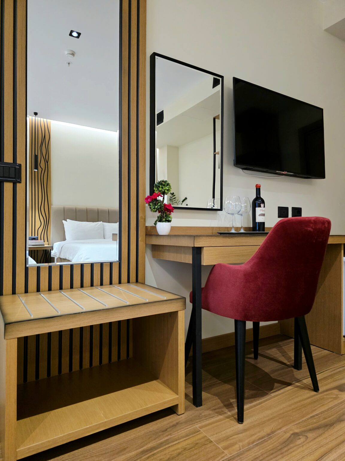 Desk corner with red velvet armchair, wine bottle and glasses, wall-mounted TV, mirror and wooden bench