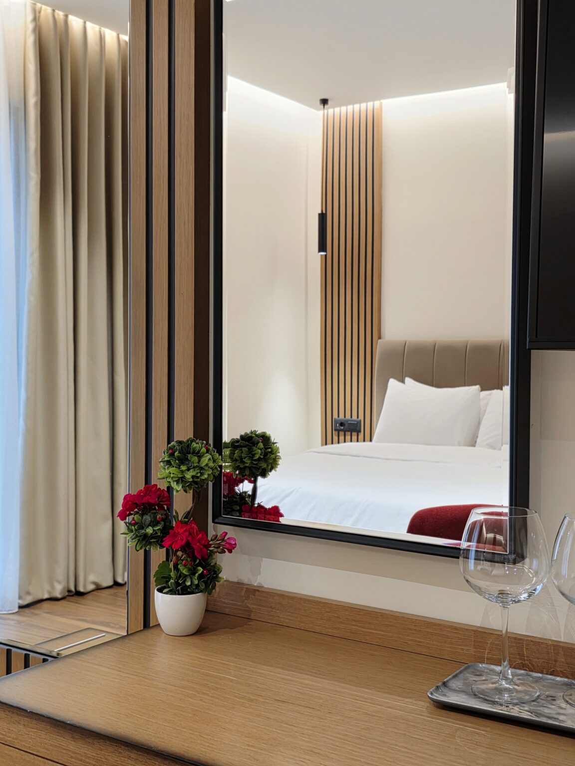 Mirror reflecting a bed and beige headboard, red flowers and wine glasses in the foreground