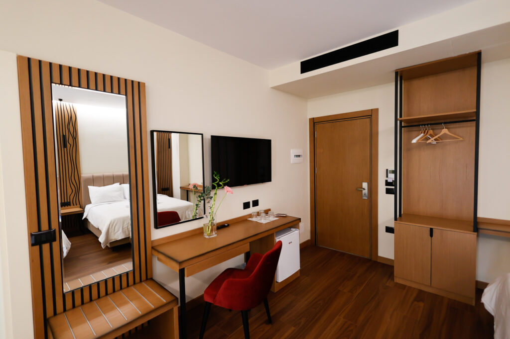Entrance area with wooden door, open wardrobe, desk, TV, red chair, minibar fridge and slatted mirror