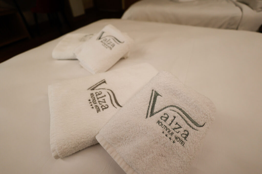 Four white towels with embroidered Valza Boutique Hotel logo, folded on a bed in Vlorë