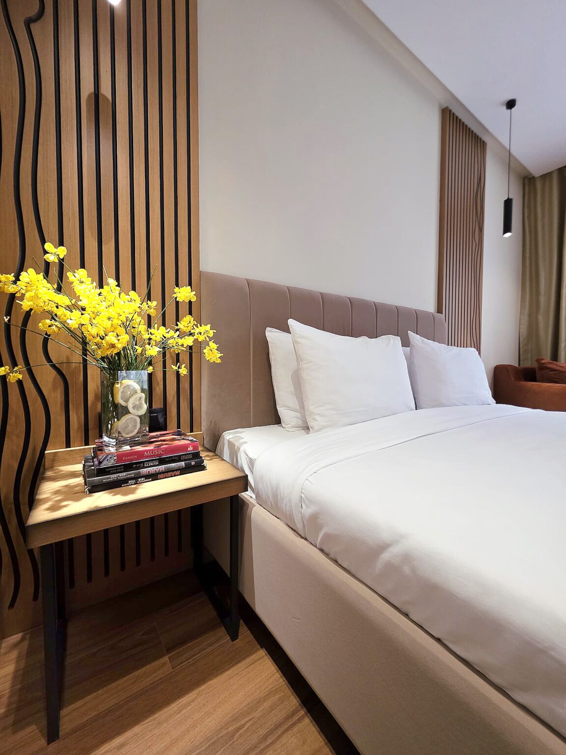 King-size bed with taupe velvet headboard, yellow flowers and books on the wooden bedside table