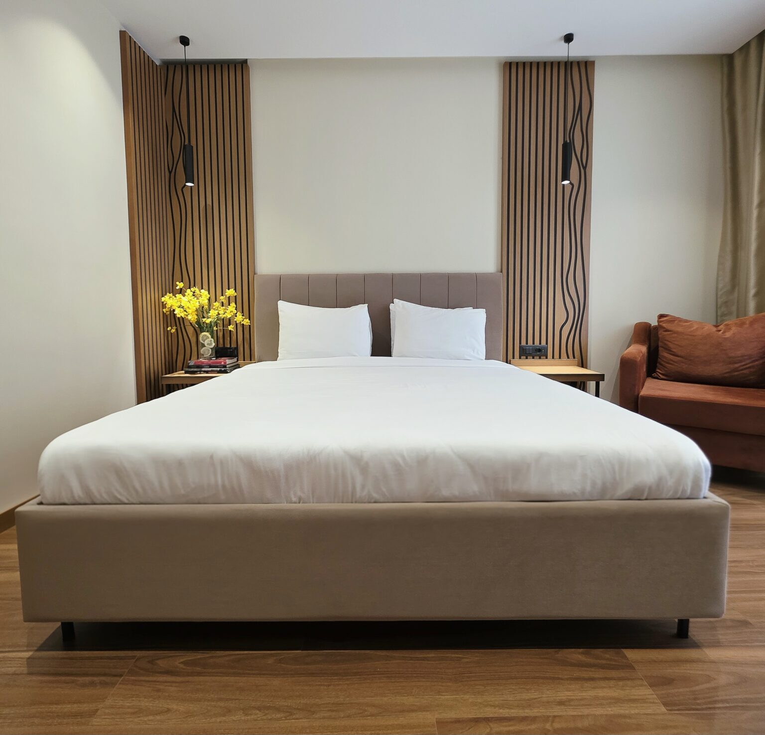 King-size bed seen from the foot, slatted wood panels with pendant lights and a rust sofa