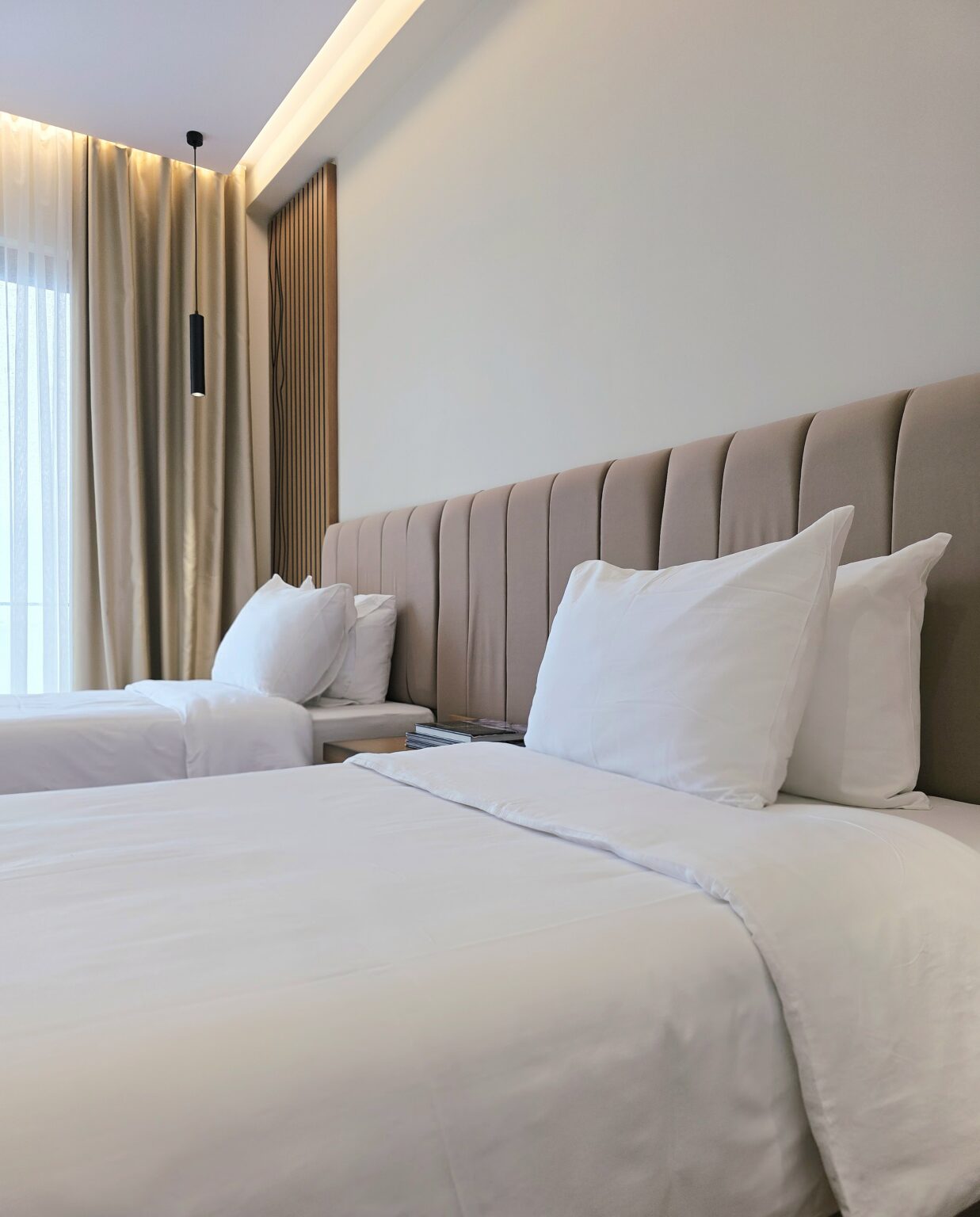 White pillows on two single beds against the beige channel-stitched headboard, black pendant lamp by the window