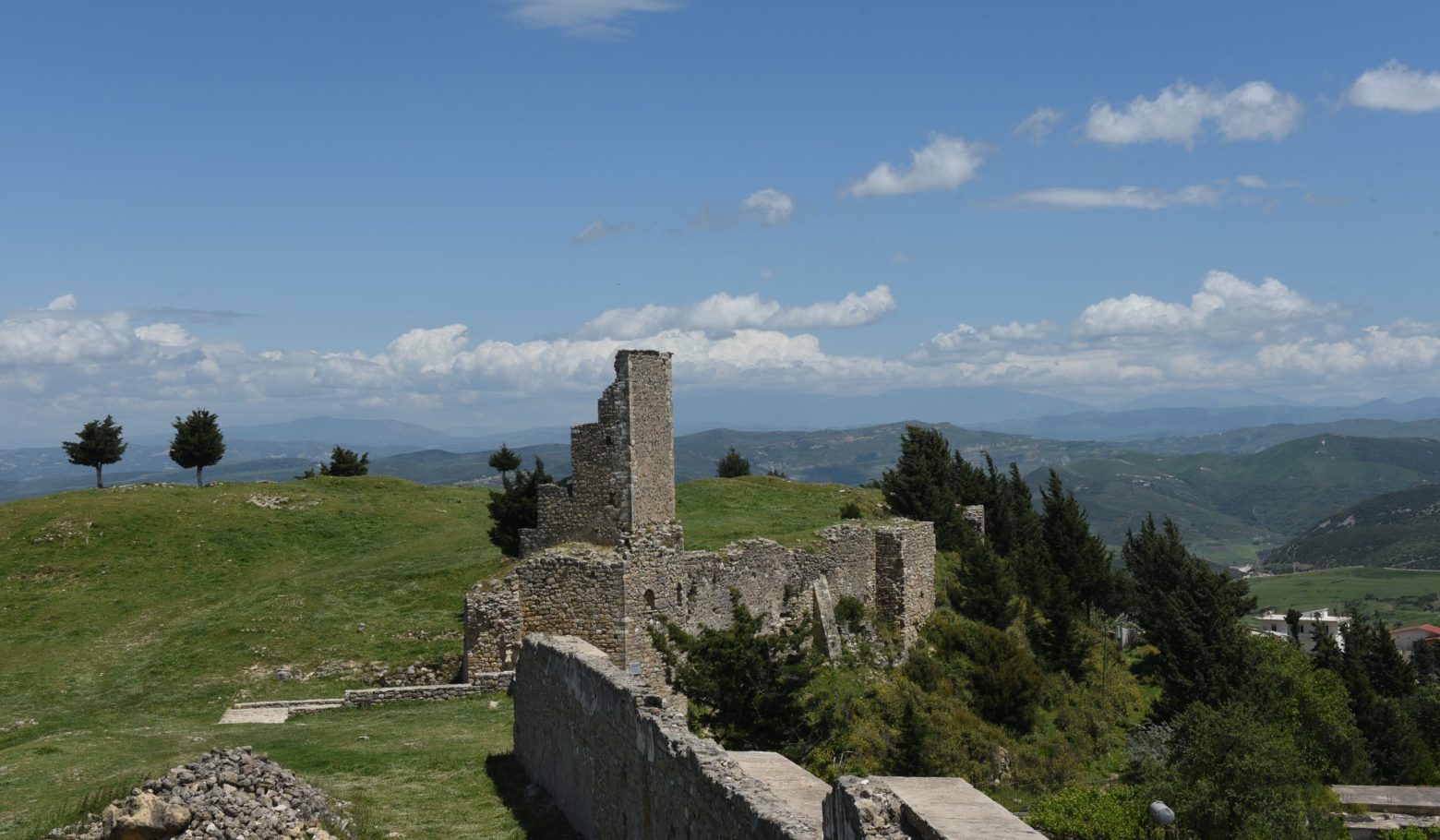 Ruined stone walls and tower of Kaninë Castle on a grassy hilltop with mountains behind