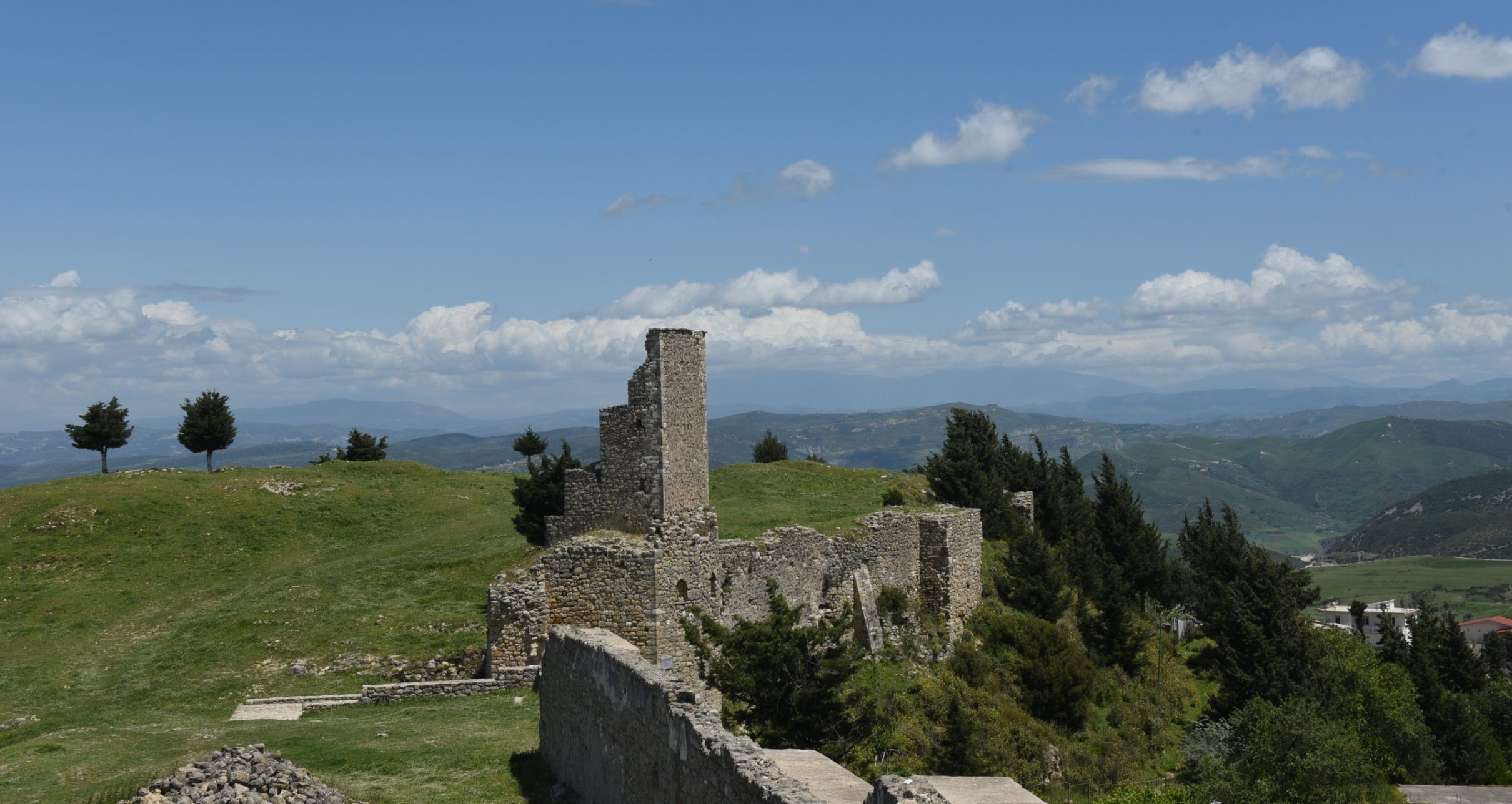 Ruined stone walls and tower of Kaninë Castle on a grassy hilltop with mountains behind