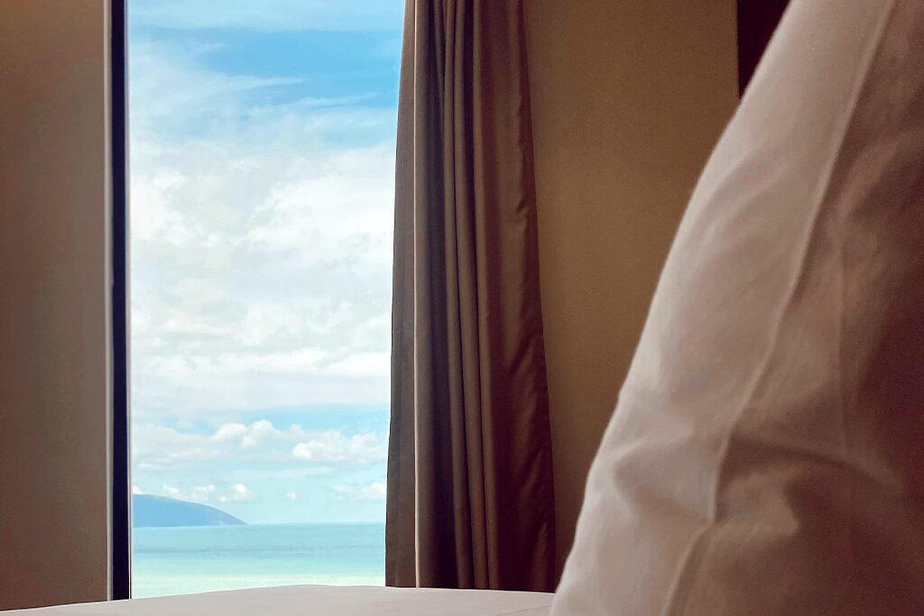 Turquoise sea and a distant headland from the Family Suite window, with a pillow and curtain in the foreground
