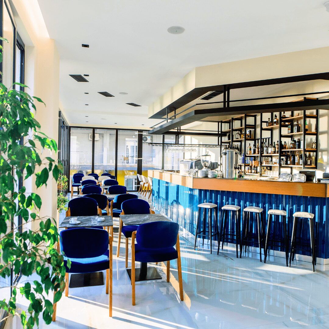 Sunlit hotel bar with blue velvet chairs, marble-topped tables and a ribbed blue counter lined with bar stools