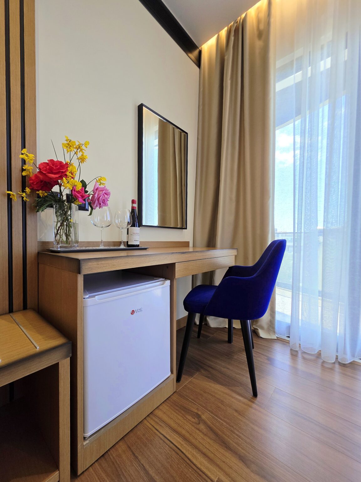 Wooden desk with a minibar underneath, blue velvet armchair and fresh flowers beside the curtained window