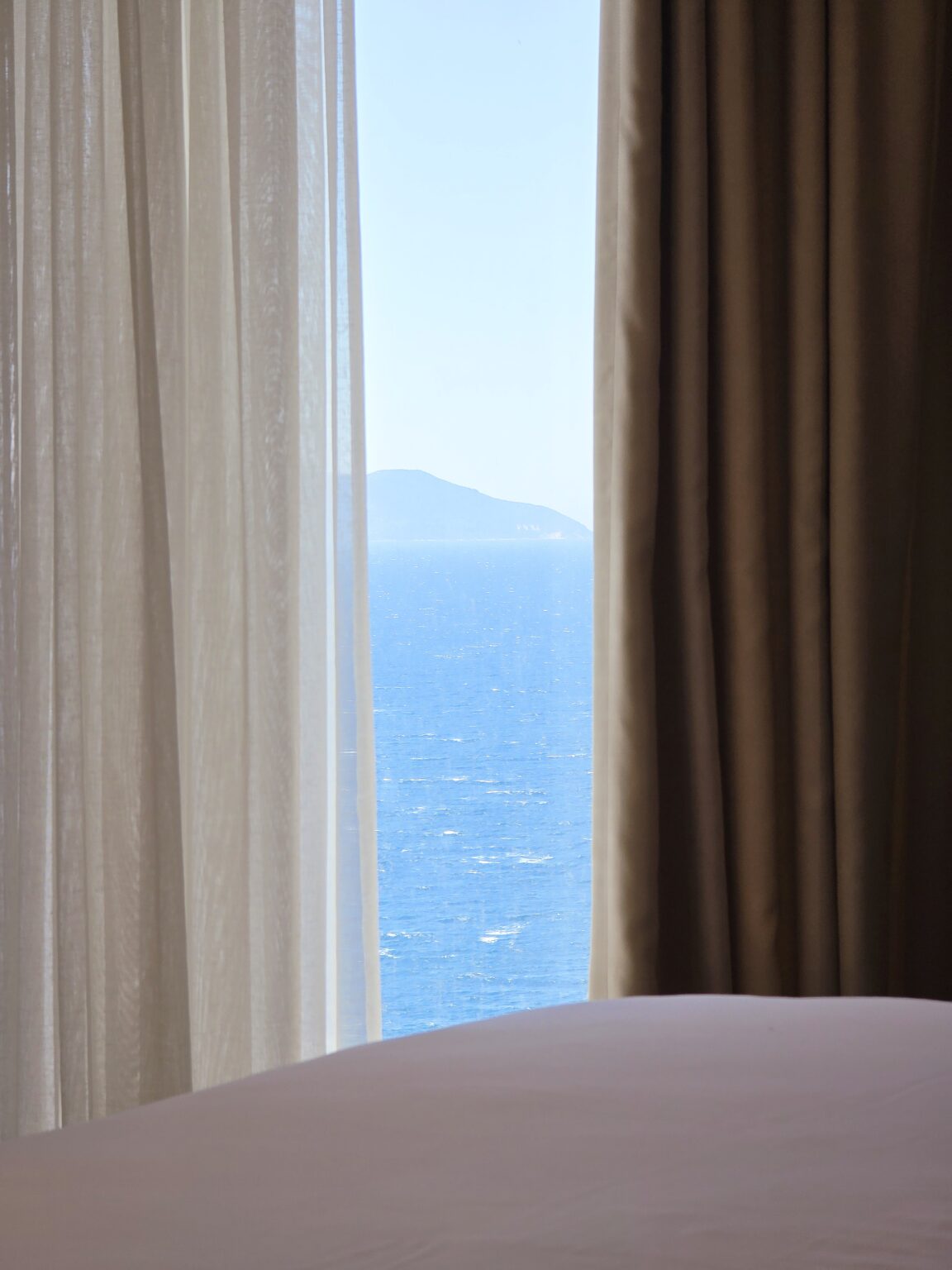Glimpse of the blue sea and a hilly headland between sheer and beige curtains in the suite