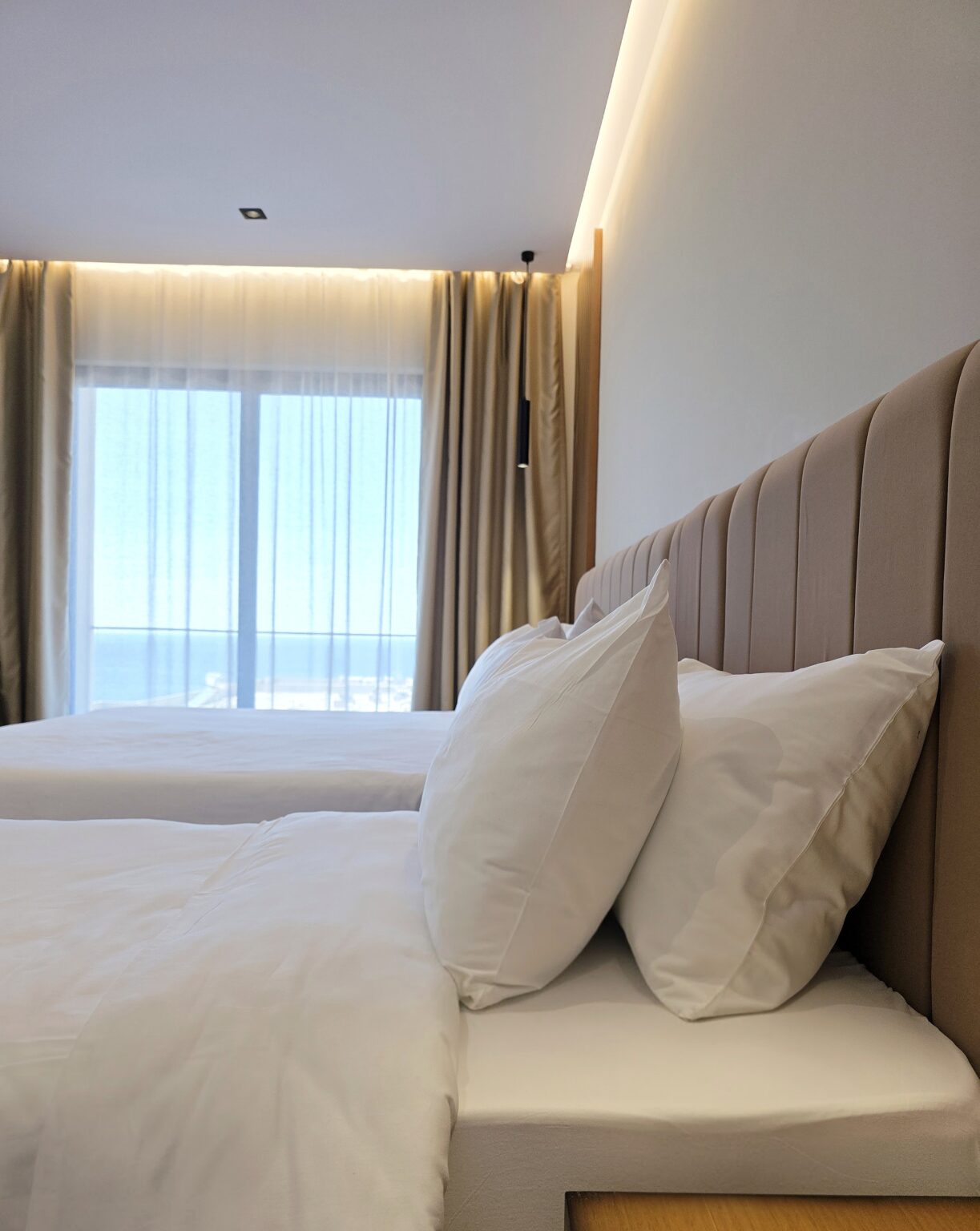 White pillows on a taupe padded headboard, with the sea at Vlorë visible through sheer curtains