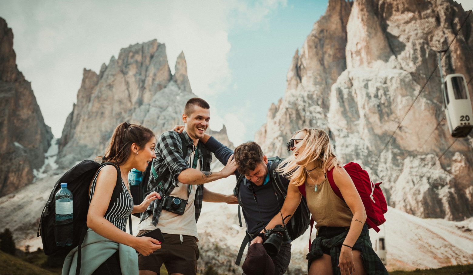 Four young hikers with backpacks laughing together below jagged rocky mountain peaks