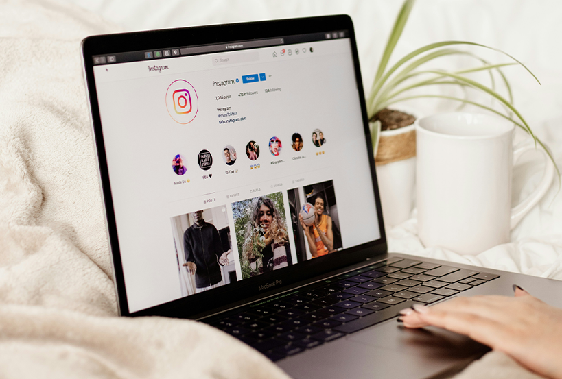 collab Laptop on a cream blanket showing an Instagram profile page, with a white mug and a plant beside it