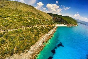 Aerial view of a green scrub-covered hillside dropping to a small white beach and turquoise cove