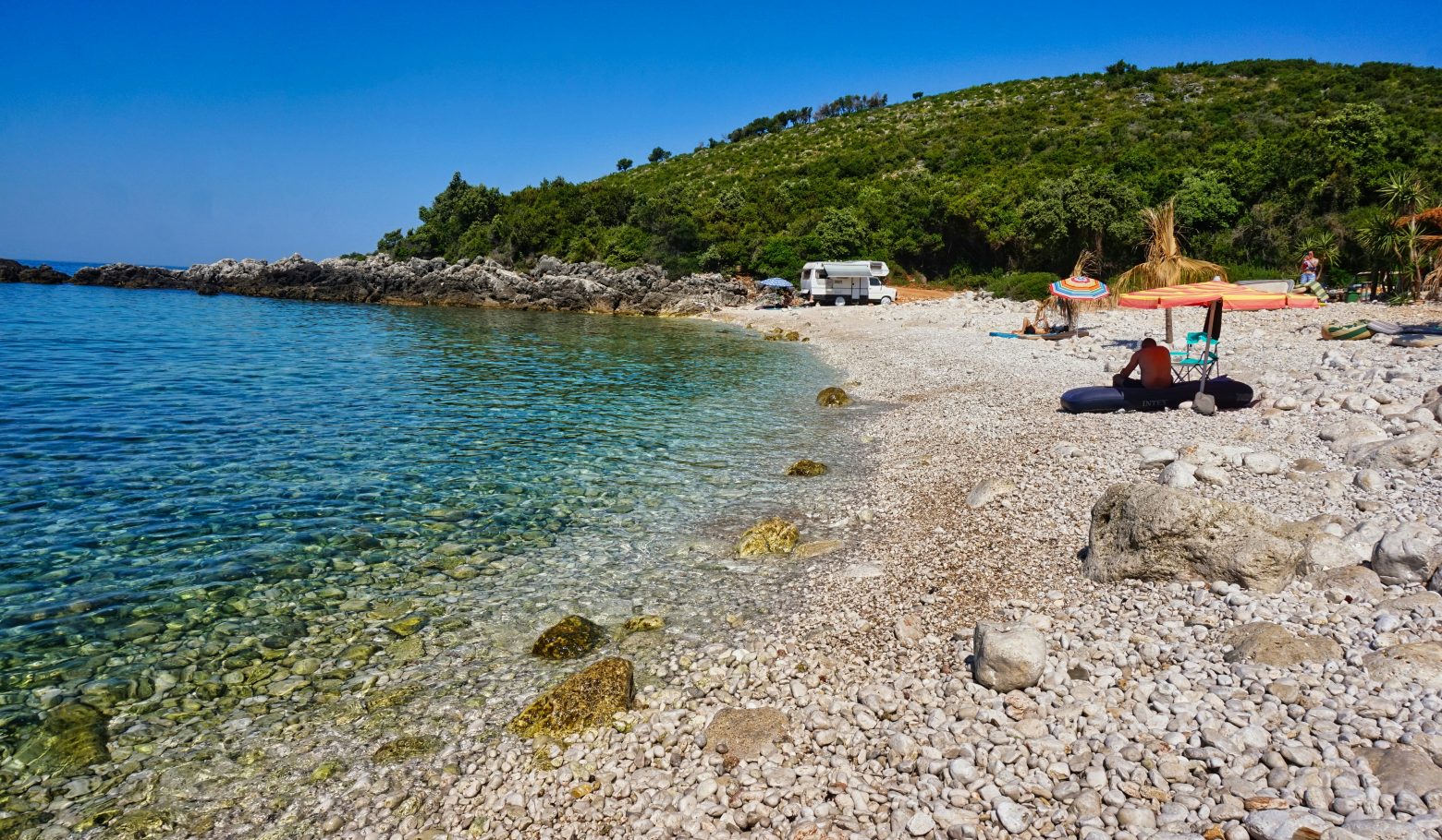 Pebble cove with clear turquoise water, a camper van and beach umbrellas below a green hillside