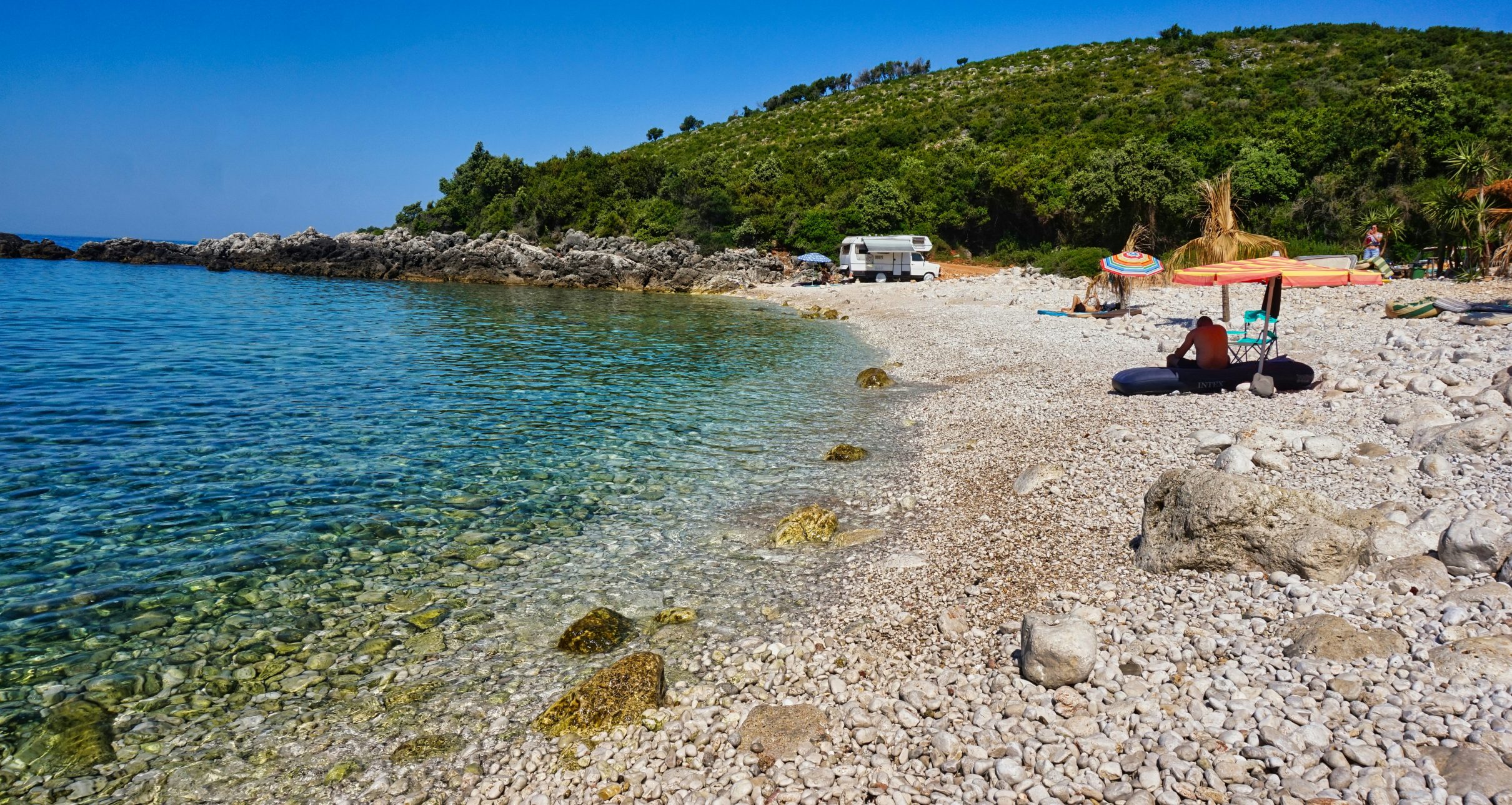 Pebble cove with clear turquoise water, a camper van and beach umbrellas below a green hillside