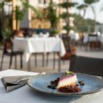 EdenRooftopRestaurant-Valza-03 Slice of berry-glazed cheesecake on a blue plate, with terrace tables in white linen behind