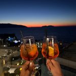 EdenRooftopRestaurant-Valza-09 Two hands raising spritz cocktails against a twilight sky over the sea, with town lights below
