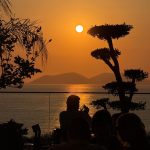 EdenRooftopRestaurant-Valza-11 Silhouetted diners and a sculpted tree against an orange sunset over the sea and distant hills