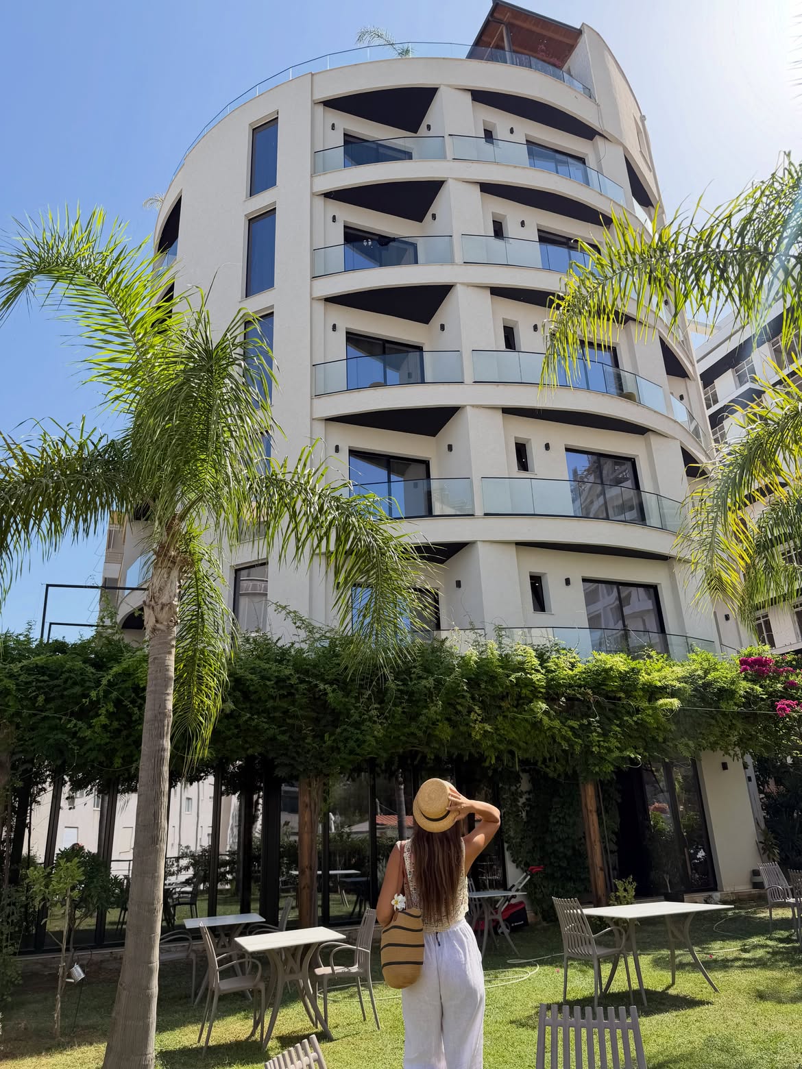 Valza Boutique Hotel curved white facade with glass balconies above a garden with palms, vines and outdoor tables