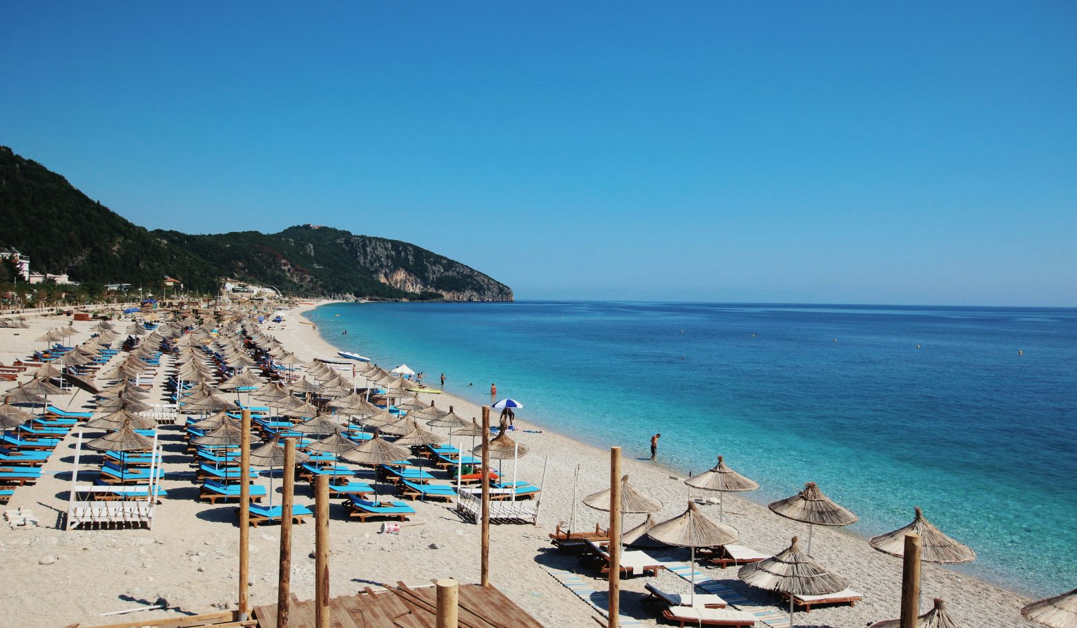 Rows of straw umbrellas and blue sun loungers on a pebble beach, with a green headland and calm sea