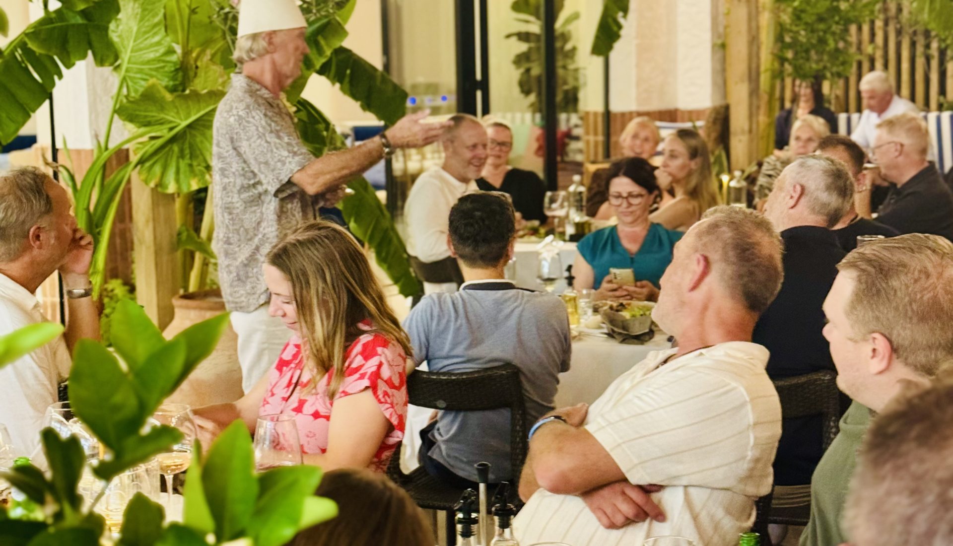 Guests at dinner tables on the terrace of Eden by Valza, under wooden beams and string lights, among olive trees and banana plants.