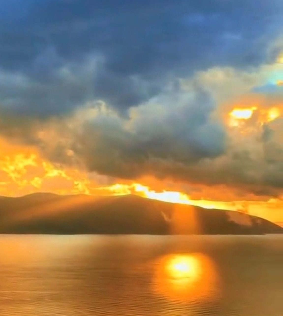 Sunset over the Bay of Vlora, with golden light breaking through dark clouds above the mountains and reflected on calm water.