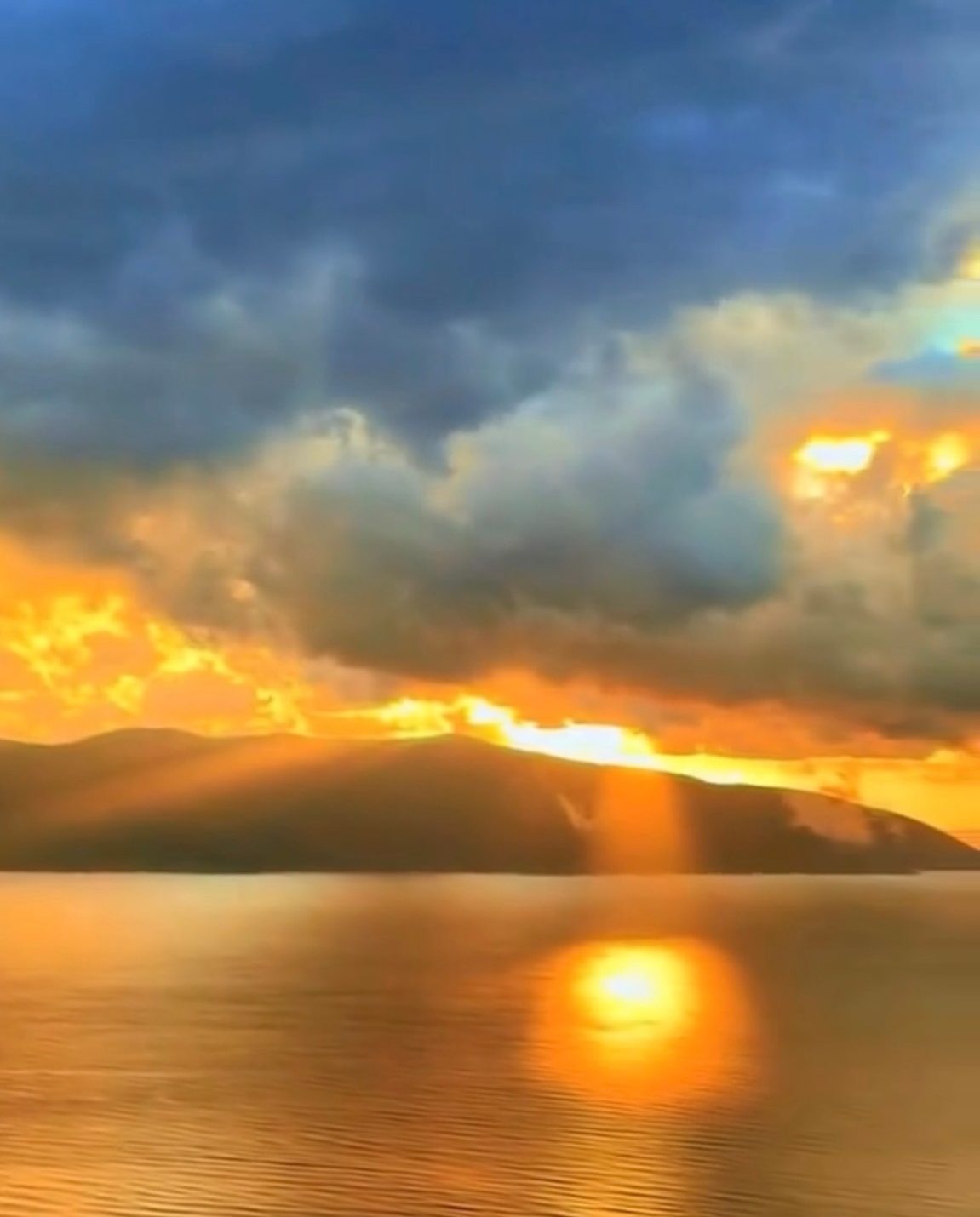 Sunset over the Bay of Vlora, with golden light breaking through dark clouds above the mountains and reflected on calm water.